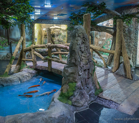 Оформление зоопарка в помещении искусственными деревьями и прудом для рыб.